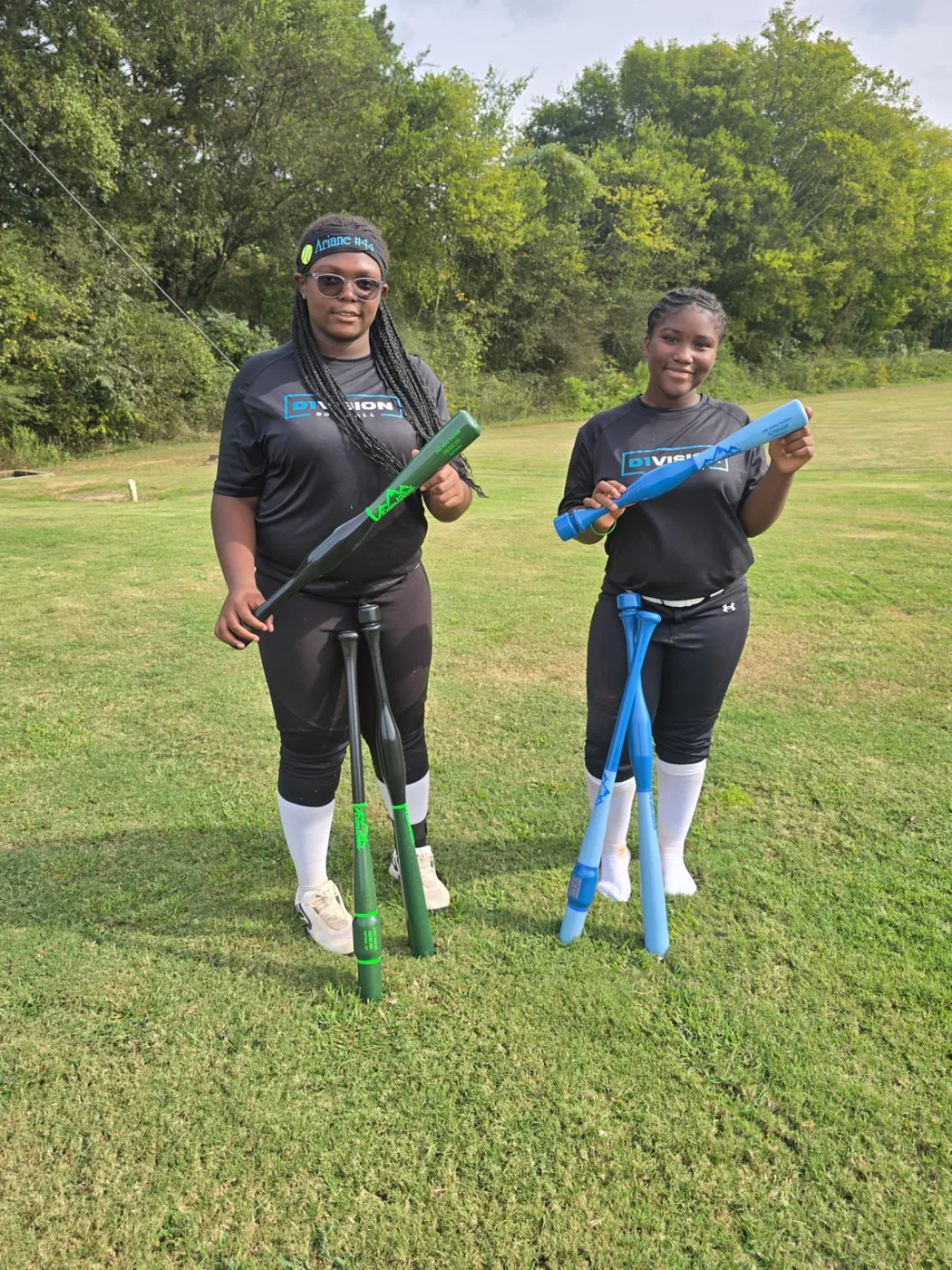 Two softball players on a grass field holding training bats